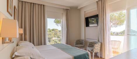 Habitación de hotel con cama, televisor, balcón con sillas y vistas a zonas verdes.