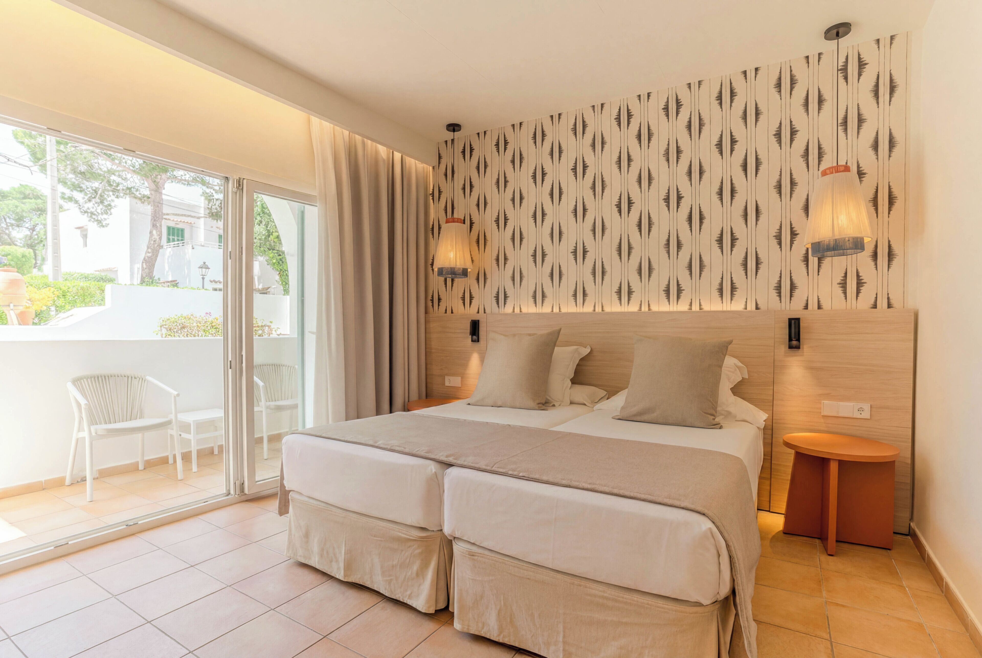 Une chambre d’hôtel avec un grand lit, deux tables de chevet, une porte-fenêtre coulissante donnant sur un espace extérieur et un mur décoré d’un motif.