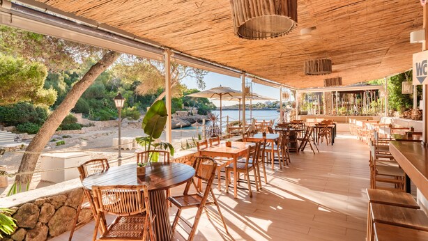 Un espace de restauration en plein air avec des tables et des chaises en bois, un toit de chaume et une vue sur la mer.