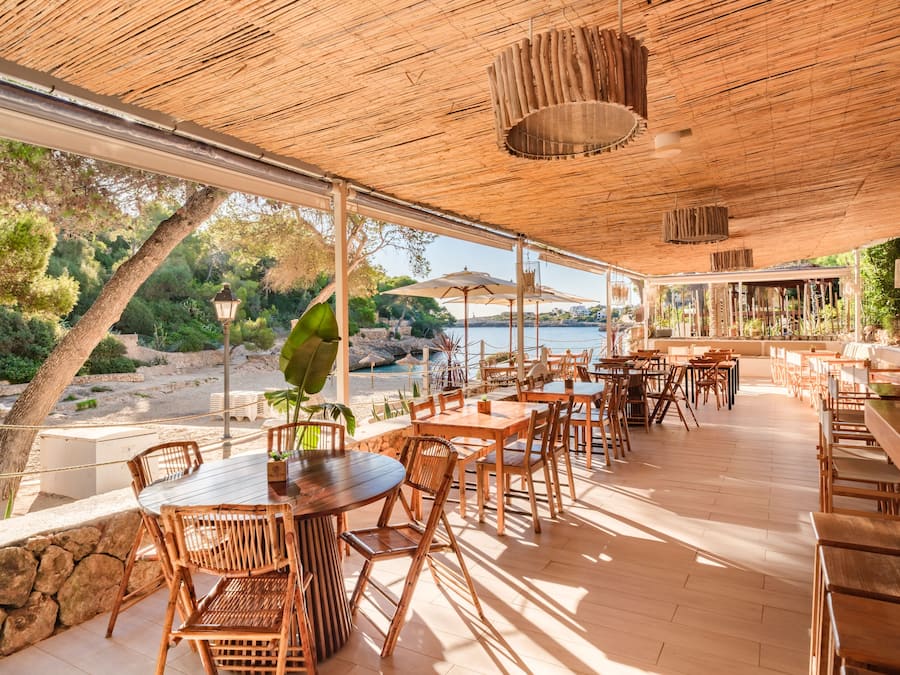 Un área de comedor al aire libre con mesas y sillas de madera, techo de paja y vista al mar.