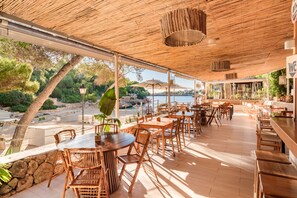 Een terras met houten tafels en stoelen, een rieten dak en uitzicht op zee.