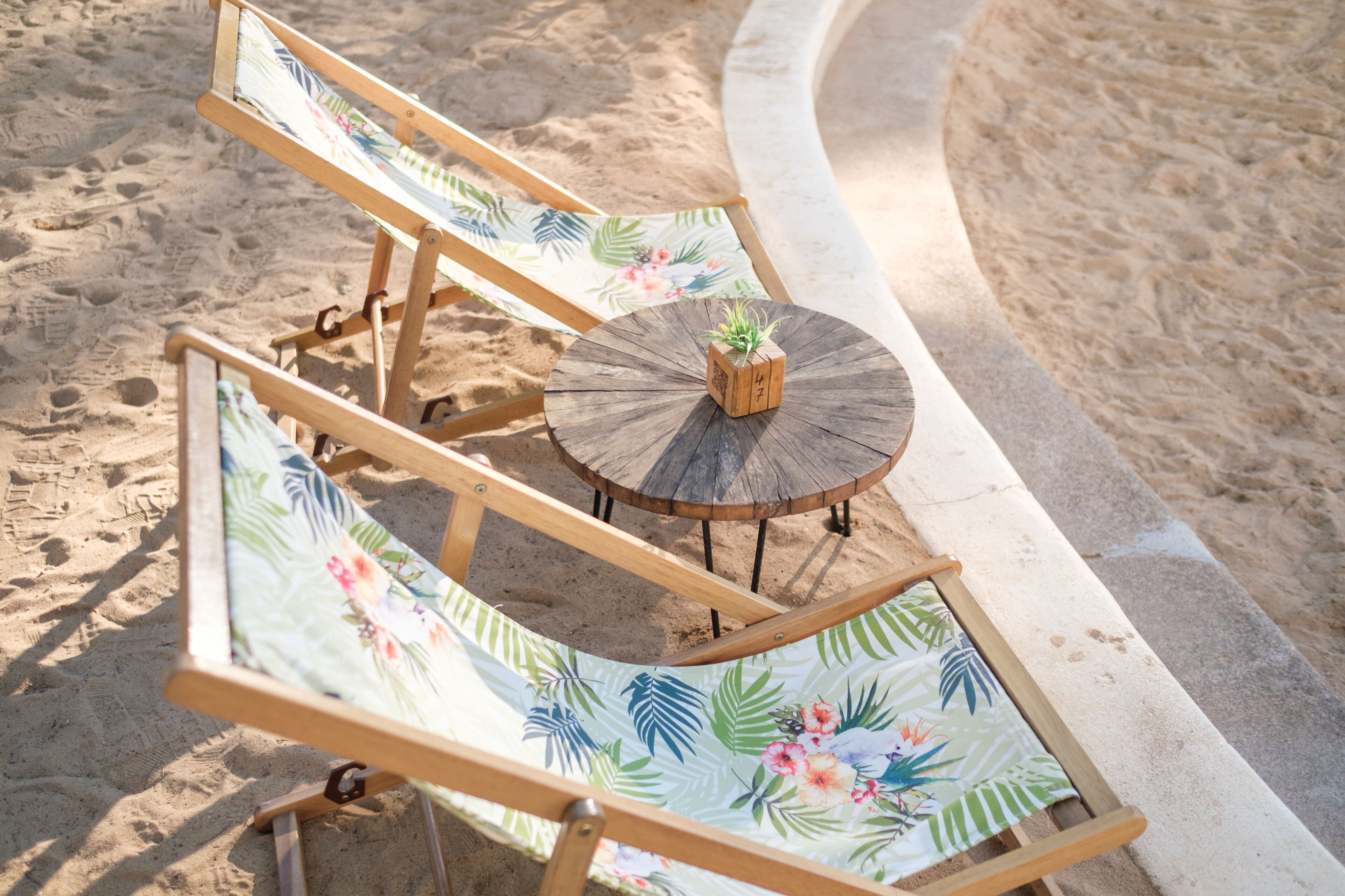 A beachside lounge with two wooden lounge chairs and a small round table, each with a floral cushion.