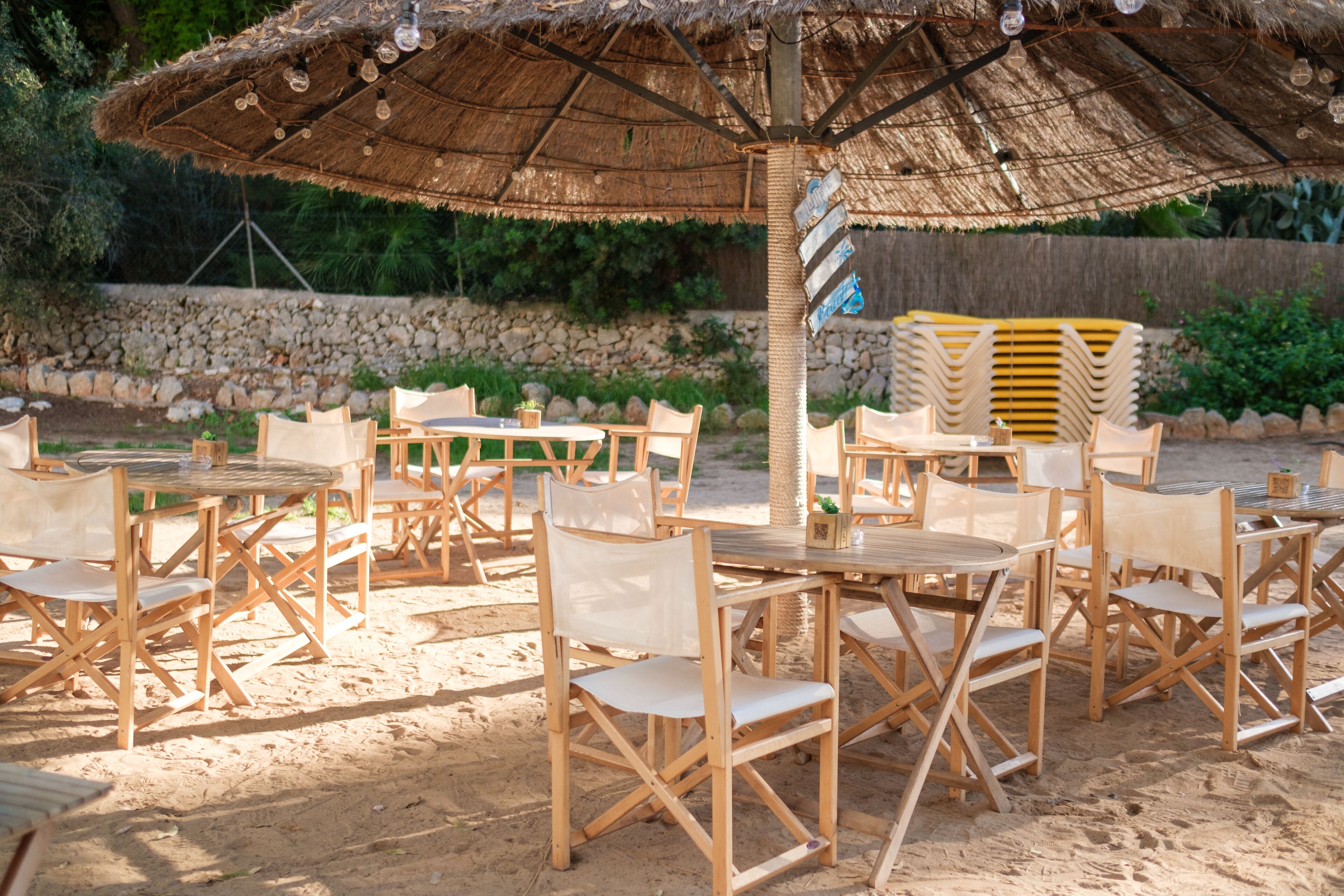An outdoor seating area with wooden tables and chairs, a thatched umbrella, and a stone wall in the background.