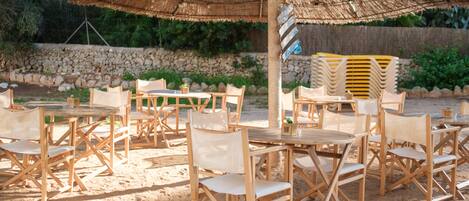 An outdoor seating area with wooden tables and chairs, a thatched umbrella, and a stone wall in the background.