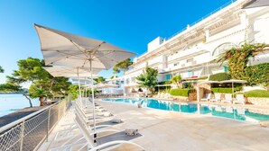 Un espace aménagé au bord de la piscine, avec des chaises longues et des parasols, une vue dégagée sur la mer, et un bâtiment doté de balcons et de verdure.