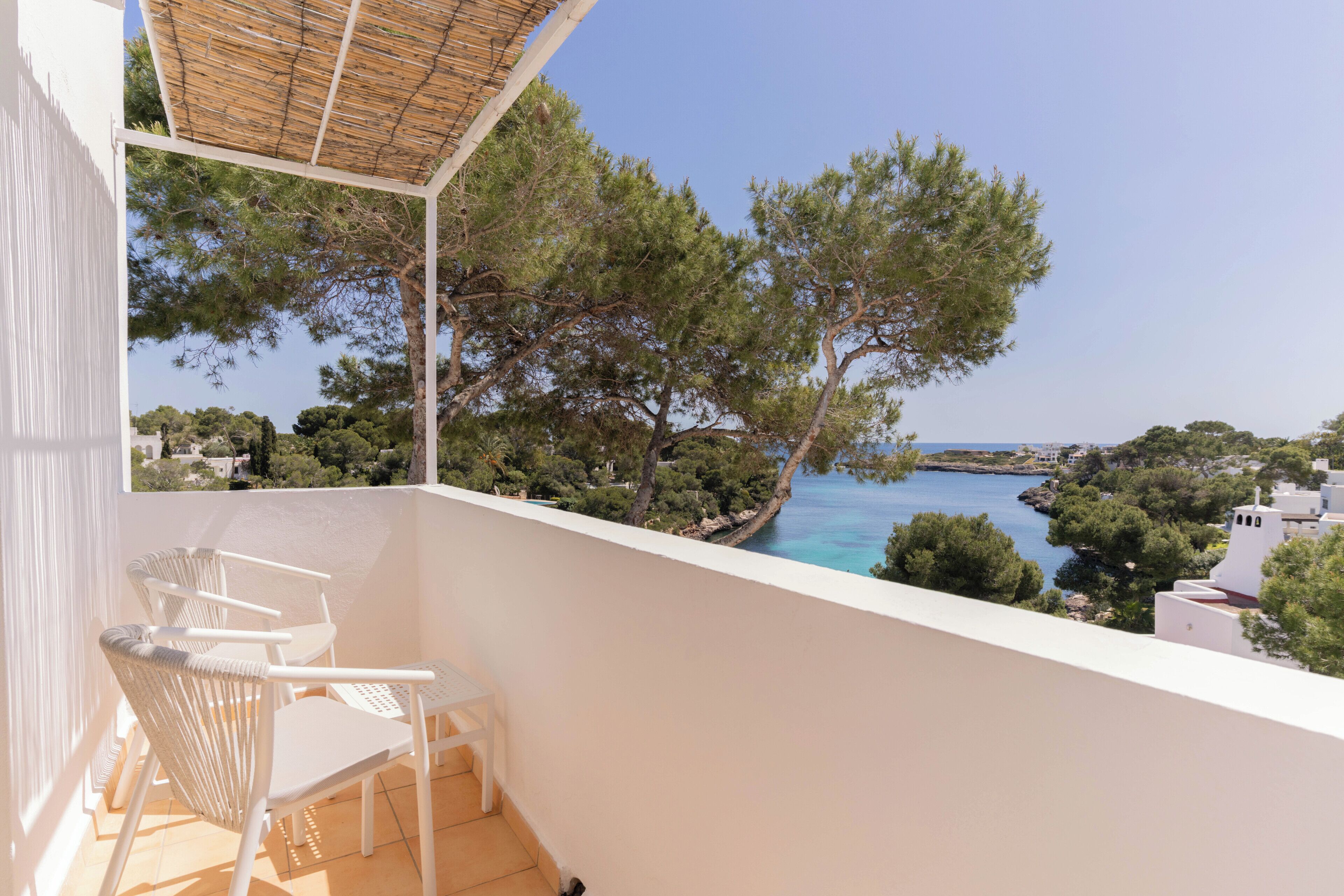 Un balcón con una silla y mesa blancos, con vistas a un paisaje costero con árboles y edificios.
