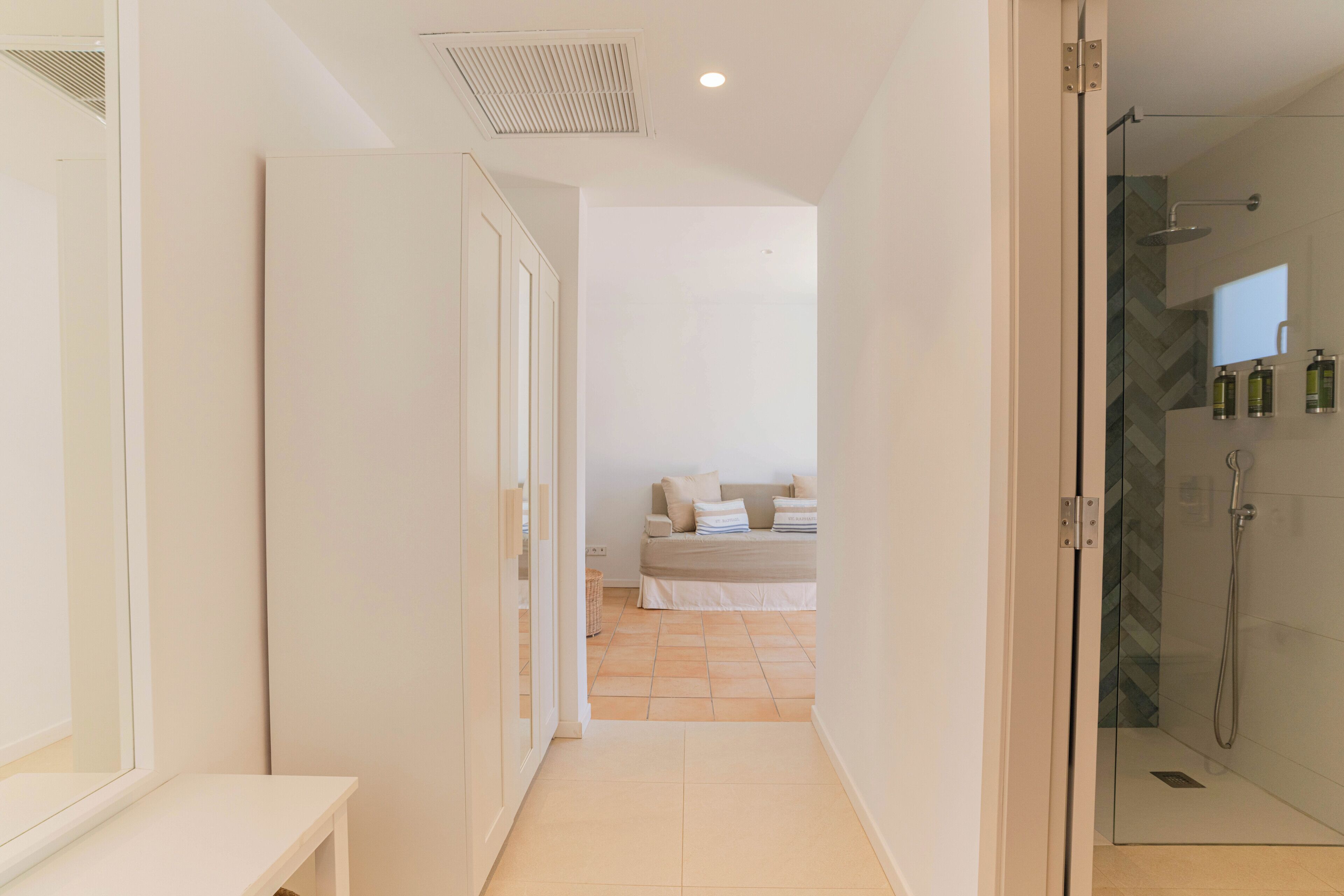 A hallway leading to a bathroom with a shower and a bed in the background.