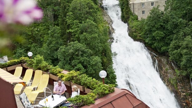 A rooftop terrace with yellow striped lounge chairs and a table, overlooking a waterfall and lush greenery. - Hotel Sanotel (Bad Gastein)