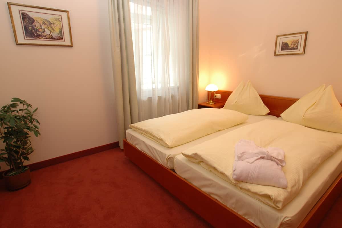 A hotel room with a bed, a nightstand, a lamp, a potted plant, and two framed pictures on the wall.
