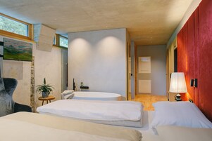 Una habitación de hotel moderna con una cama grande, una mesita de noche pequeña con lámpara, un ventanal y vistas al exterior.