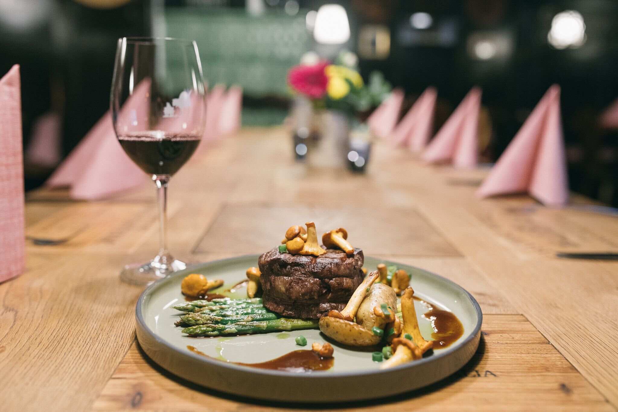 A plate of steak with asparagus and potatoes, a glass of red wine, and a vase of flowers on a wooden table.