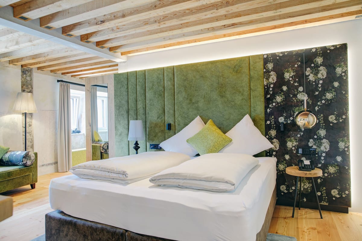 Ein modernes Schlafzimmer mit Holzboden, einem grünen gepolsterten Kopfteil, einem Bett mit weißen Bettbezügen, einem kleinen Nachttisch mit Lampe und floralem Tapetenmuster an der Wand.