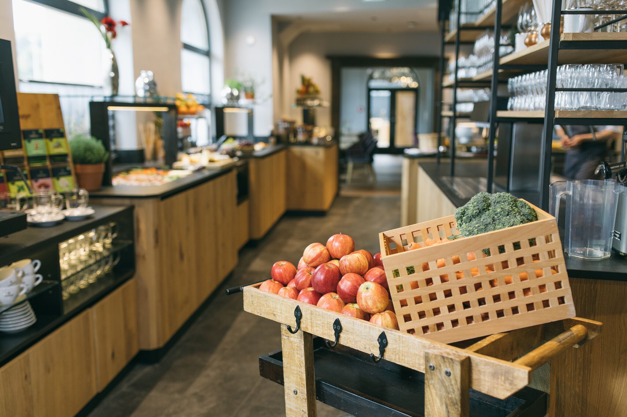 A kitchen area with wooden cabinets, a fruit display with apples and broccoli, and a coffee station with cups and a coffee machine.
