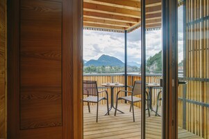 Una terraza de madera con un ventanal de vidrio que da a un balcón con mesa y sillas, con vistas a un paisaje montañoso.