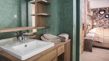 A bathroom with a wooden vanity, a white sink, and a mirror. There are towels stacked on the vanity and a shelf above the mirror.
