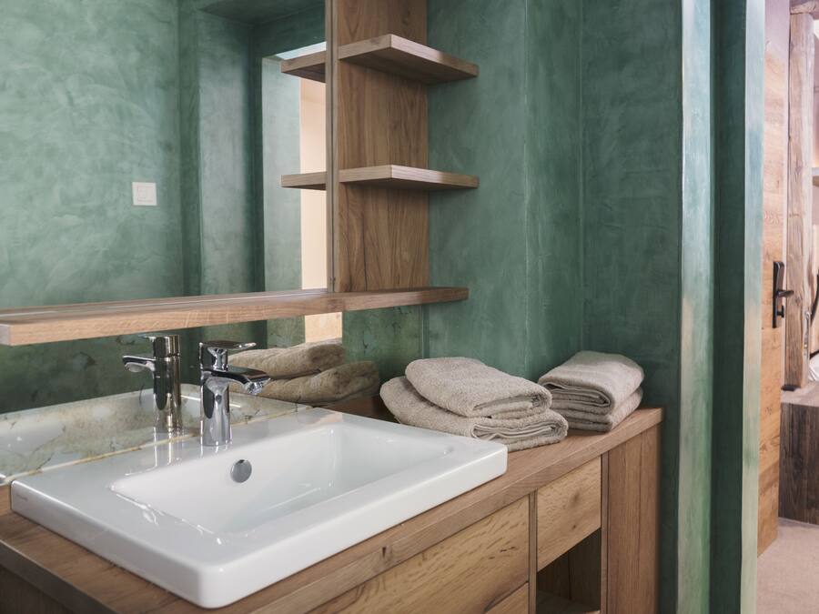 A bathroom with a wooden vanity, a white sink, and a mirror. There are towels stacked on the vanity and a shelf above the mirror.