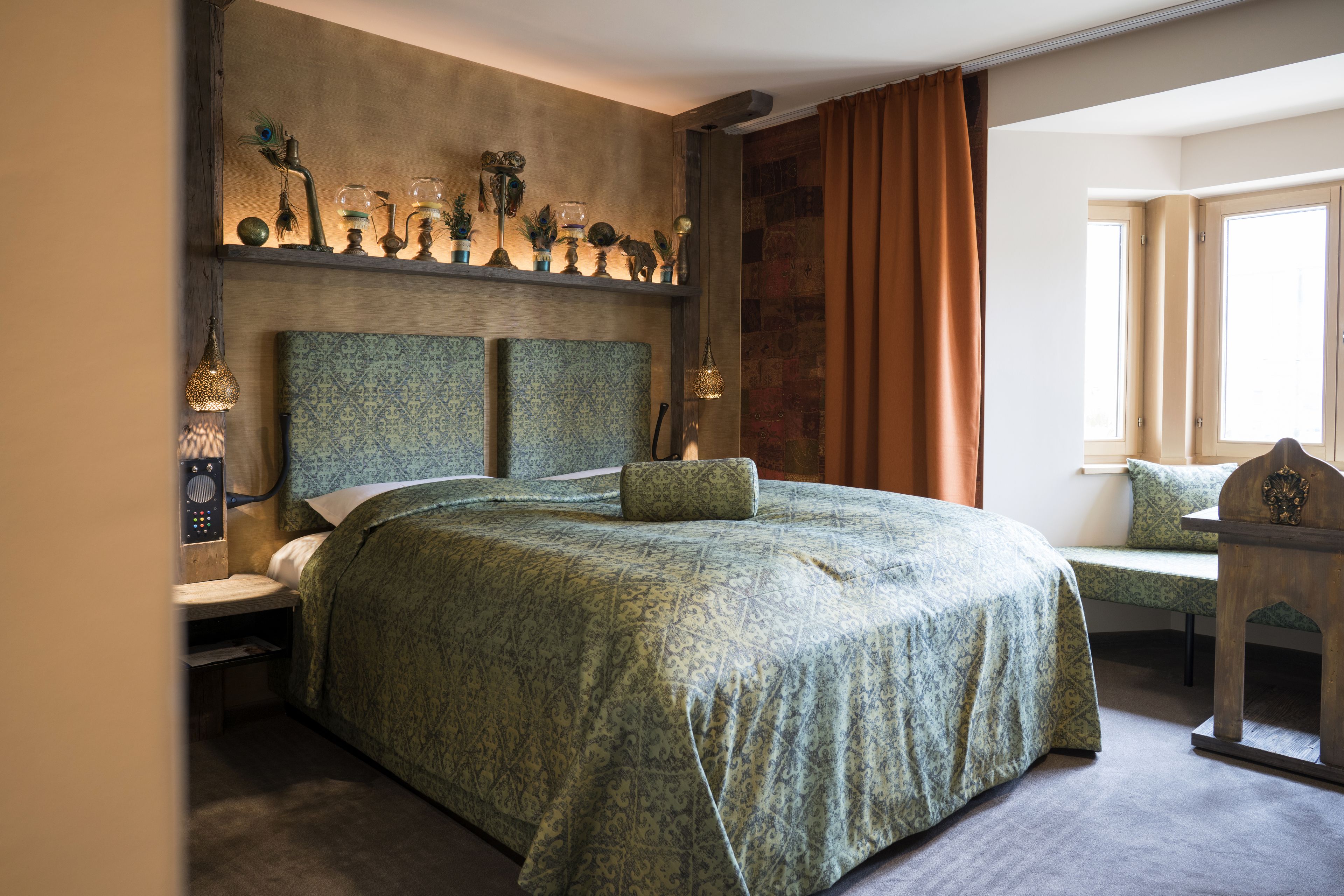 A large bed with a patterned bedspread, a bedside table with a lamp, and a decorative shelf above the bed.