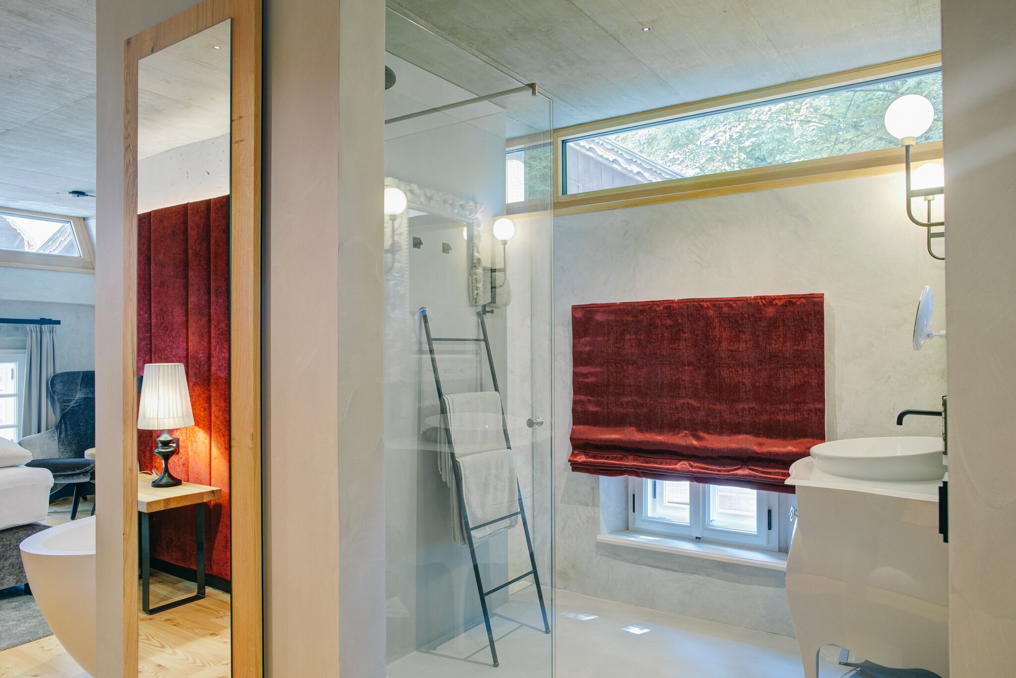 A modern bathroom with a glass shower enclosure, a red towel, a white sink, and a window with a view of greenery.