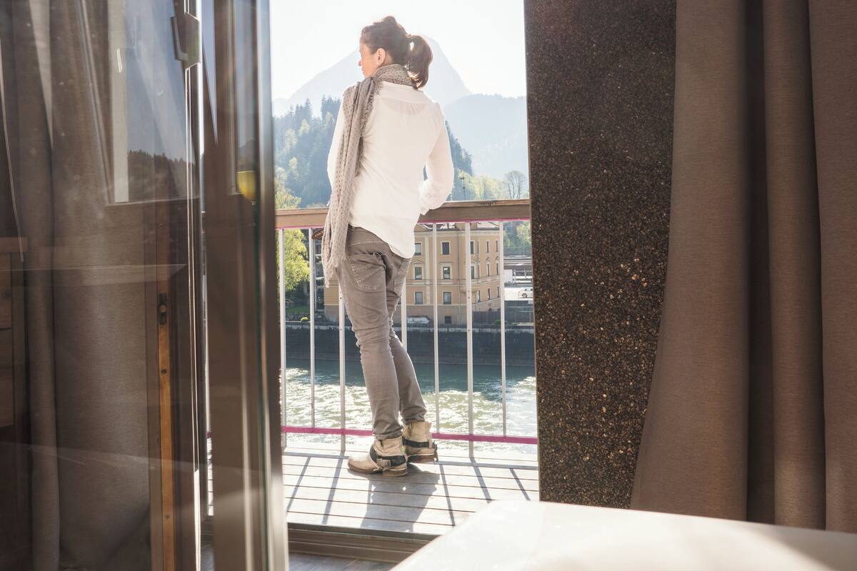 Eine Person steht vor einer Glastür mit Blick auf ein Gewässer und Gebäude im Hintergrund.