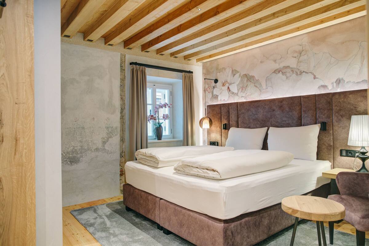 Ein Schlafzimmer mit Holzboden, einem Bett mit weißen Bettwäsche, einem Nachttisch, einer Lampe und Blick aus einem Fenster mit Vorhängen.