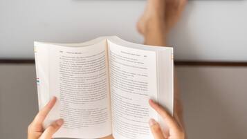 Una persona leyendo un libro en una habitación con una cama y un tapiz de pared con motivos geométricos.