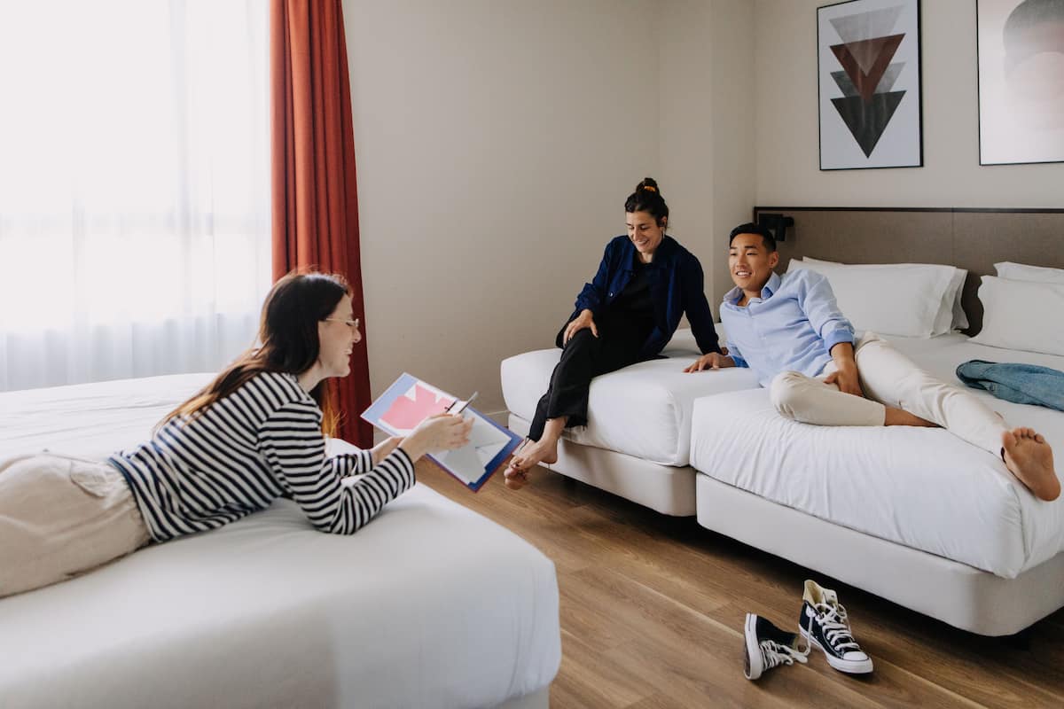 Et moderne hotelværelse med en stor seng, en sofa og et lille bord. To personer sidder på sofaen, hvoraf den ene holder en papirklemme.