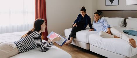 Una habitación con cama, un sofá y una mesita de centro. Dos personas están sentadas en el sofá, una sosteniendo un libro.
