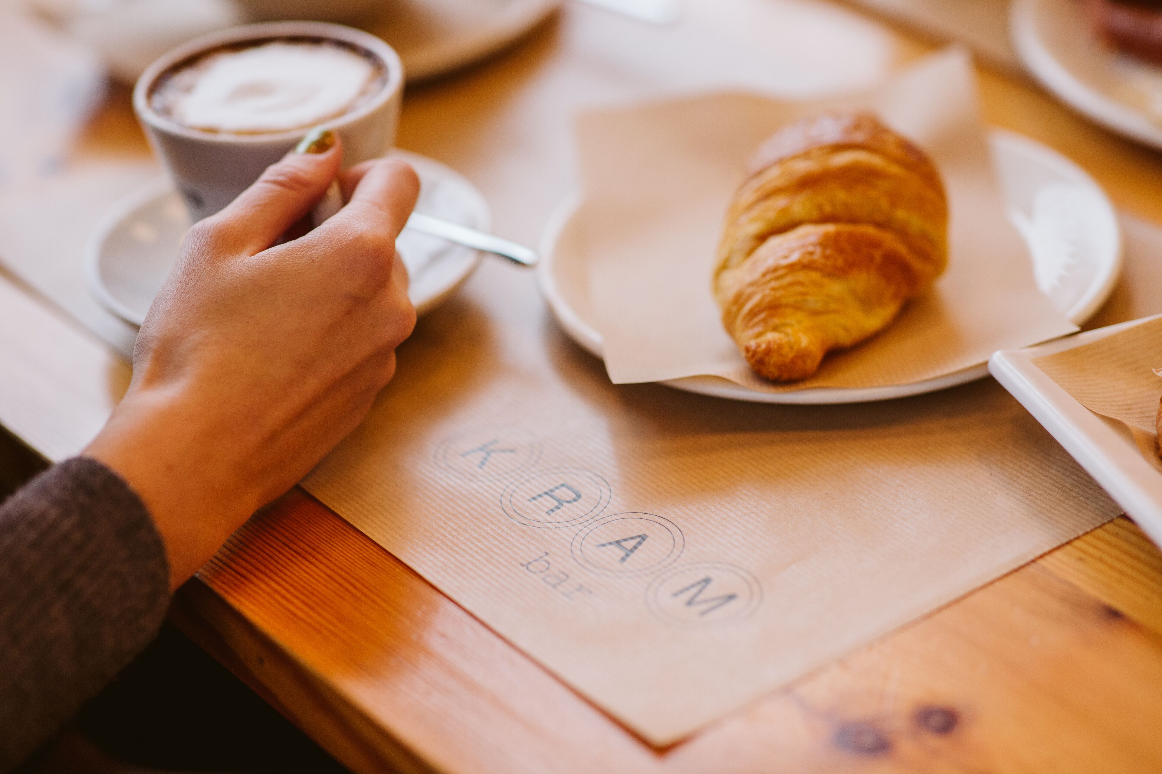 Una mano sosteniendo una cuchara sobre una taza de café, un croissant en un plato y una servilleta con el texto 'KRAM bar'.