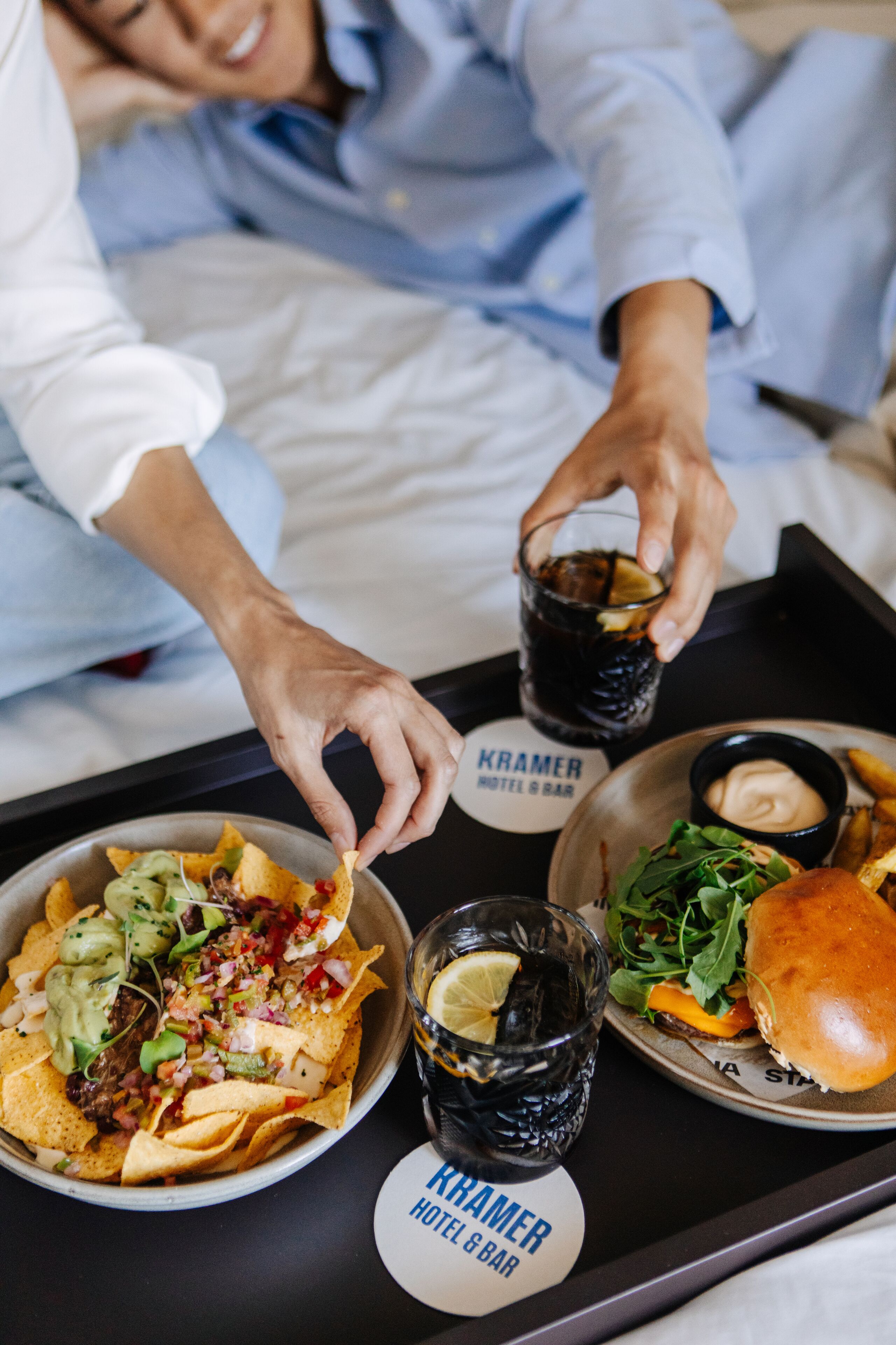 A tray with nachos, a burger, and two glasses of a dark beverage, labeled "Kramer Hotel & Bar".