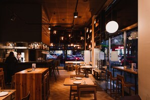 A cozy restaurant with wooden tables and chairs, a bar area, and large windows offering a view of the street outside. - Hotel Kramer (Valencia)