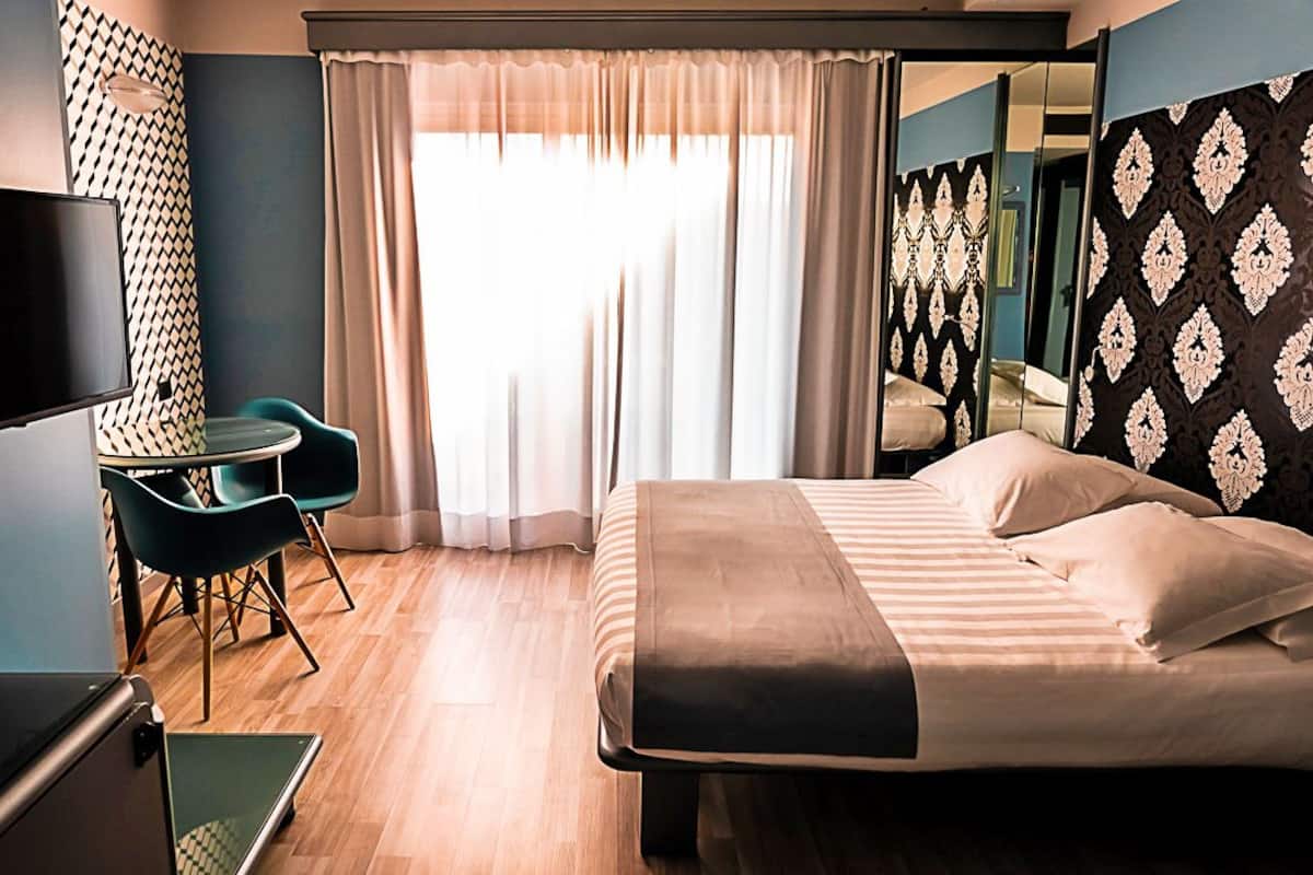 Une chambre d’hôtel avec un lit, un bureau et une chaise, une télévision et une grande fenêtre avec des rideaux.