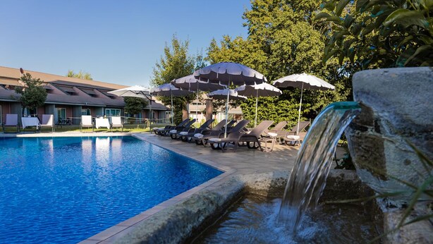 A swimming pool with lounge chairs and umbrellas, a waterfall feature, and a building with a tiled roof in the background. - Business Hotel (Casale Monferrato)