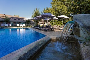 A swimming pool with lounge chairs and umbrellas, a waterfall feature, and a building with a tiled roof in the background. - Business Hotel (Casale Monferrato)