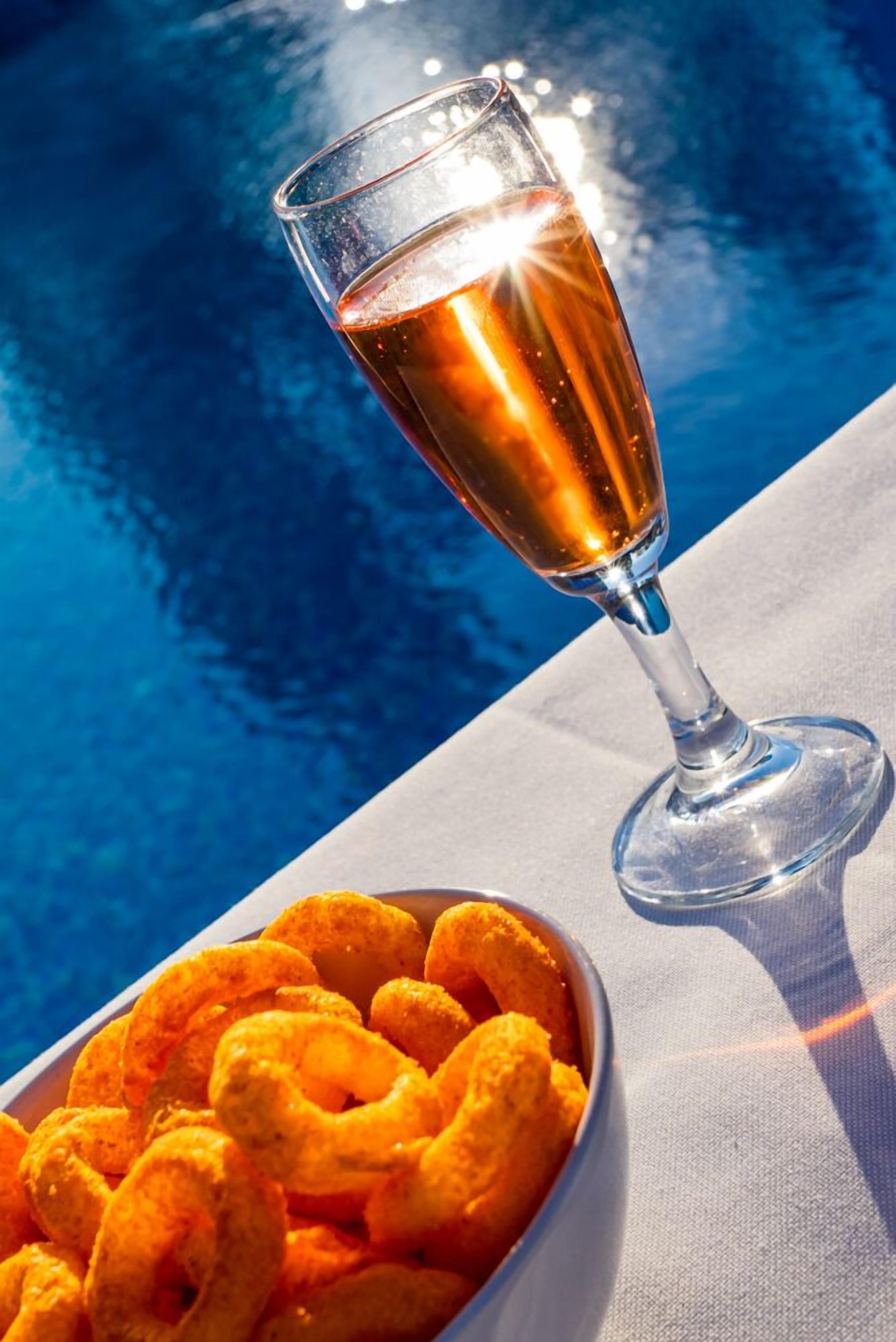 A glass of amber liquid next to a bowl of golden-brown fried snacks by a pool.