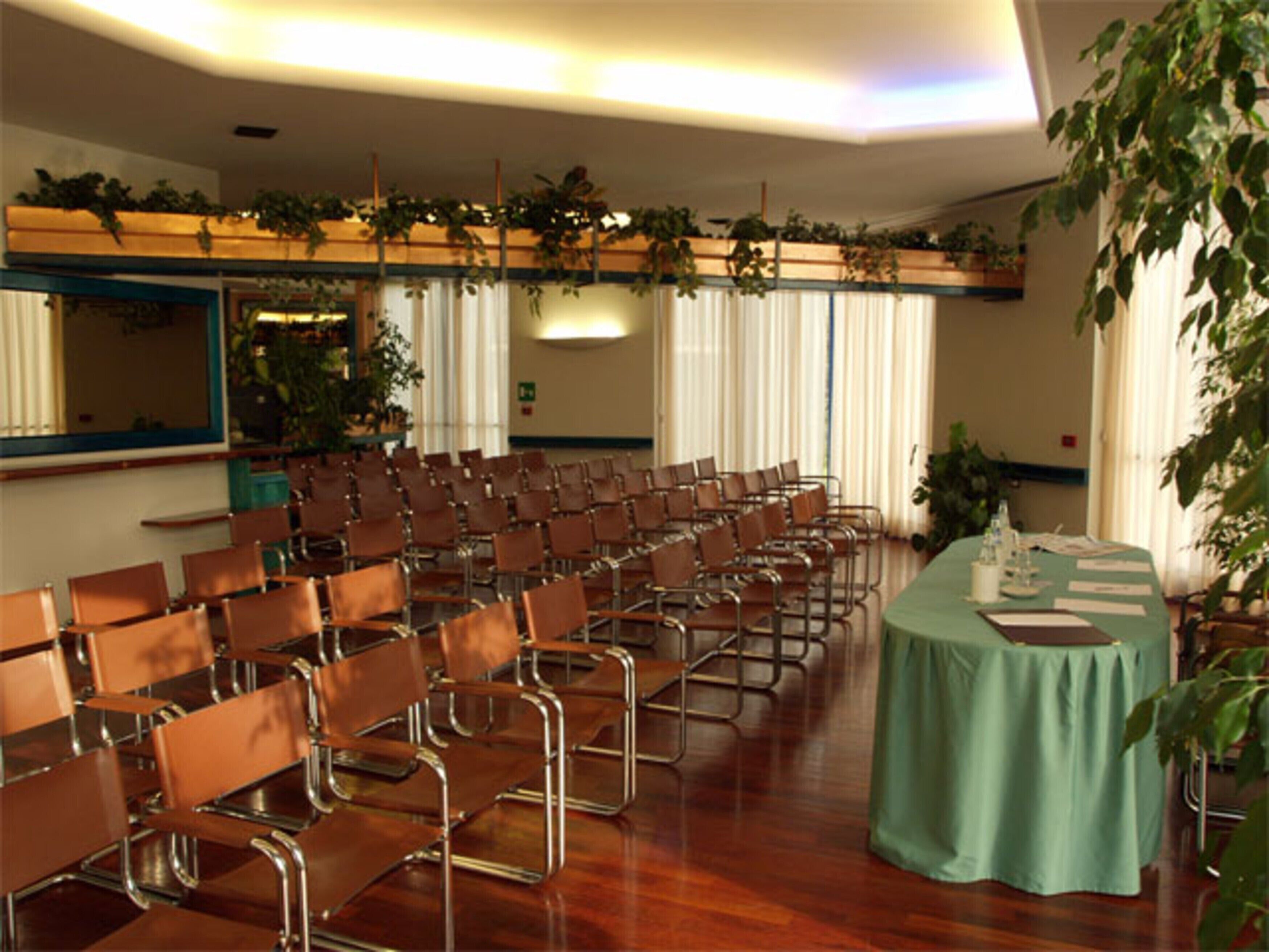 Un salón de conferencias con filas de sillas marrones, una mesa larga con mantel verde y plantas en el techo.