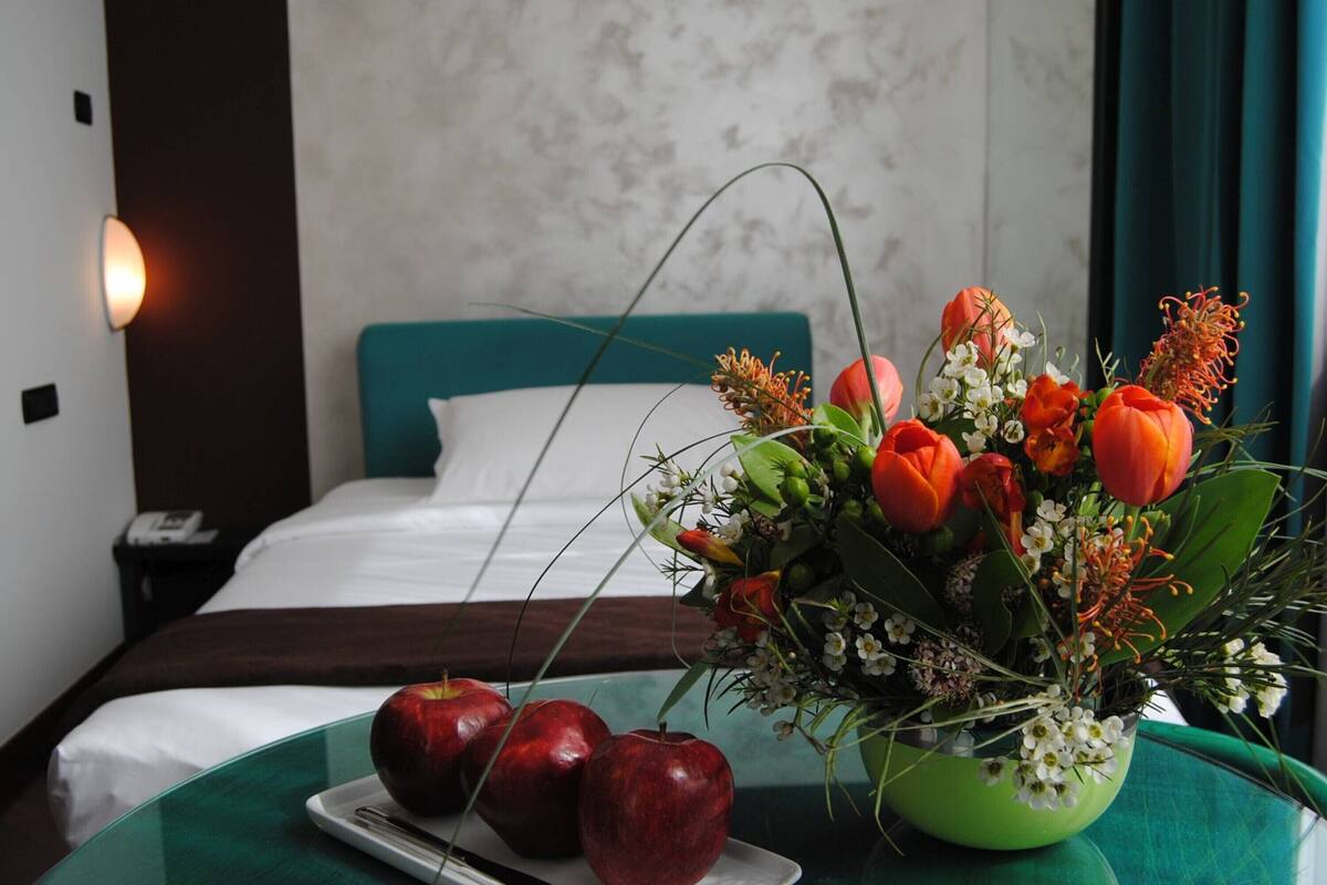 Une chambre d’hôtel avec un lit, une table de chevet avec un téléphone, une table verte avec des pommes et un arrangement floral.
