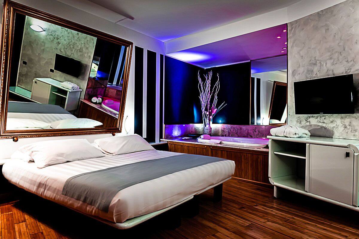Une chambre d’hôtel moderne dotée d’un grand lit, d’une coiffeuse avec un miroir, d’un coin salon avec un canapé violet et d’un téléviseur fixé au mur.