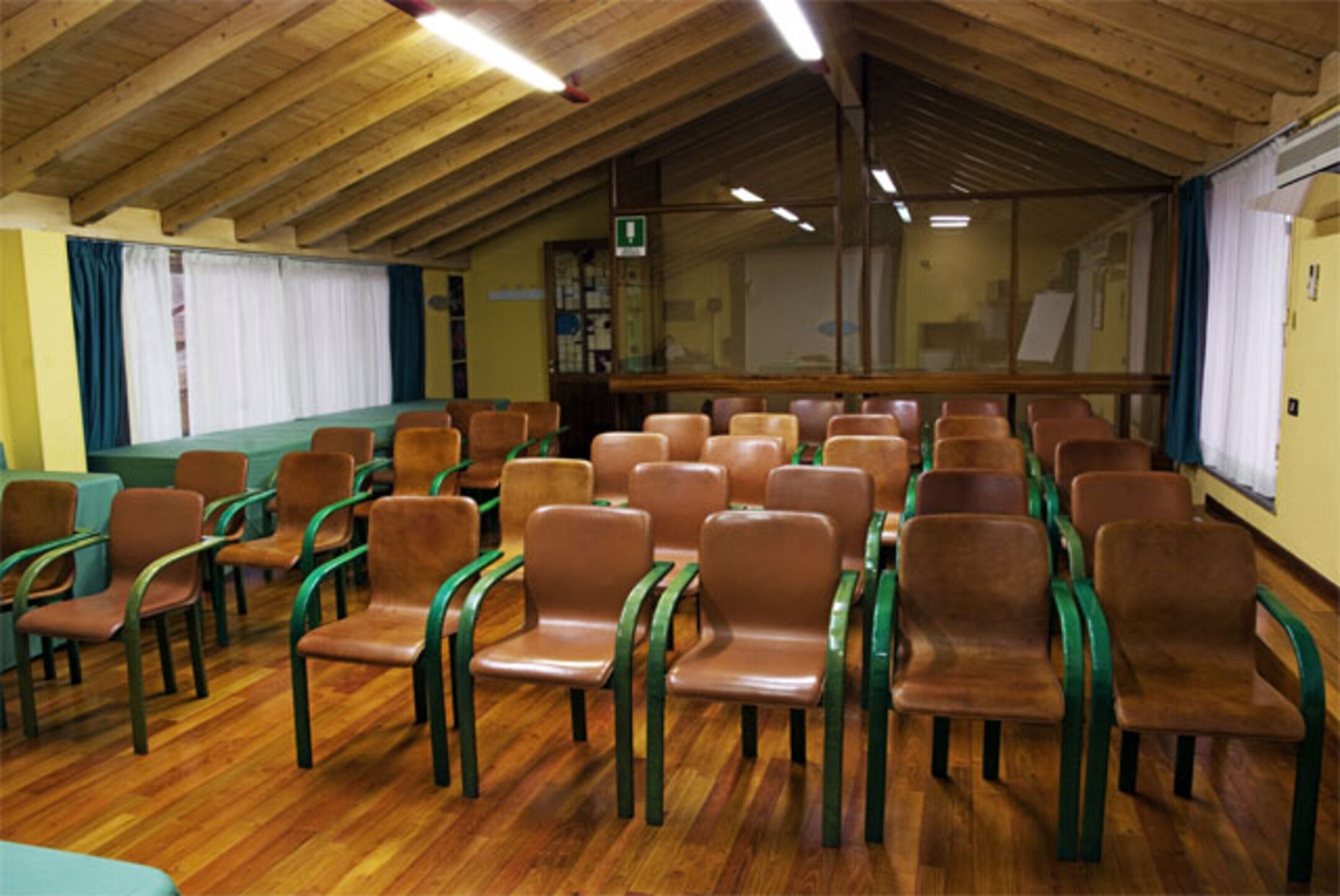 A room with rows of brown chairs and green armrests, wooden floors, and a high wooden ceiling with fluorescent lighting.
