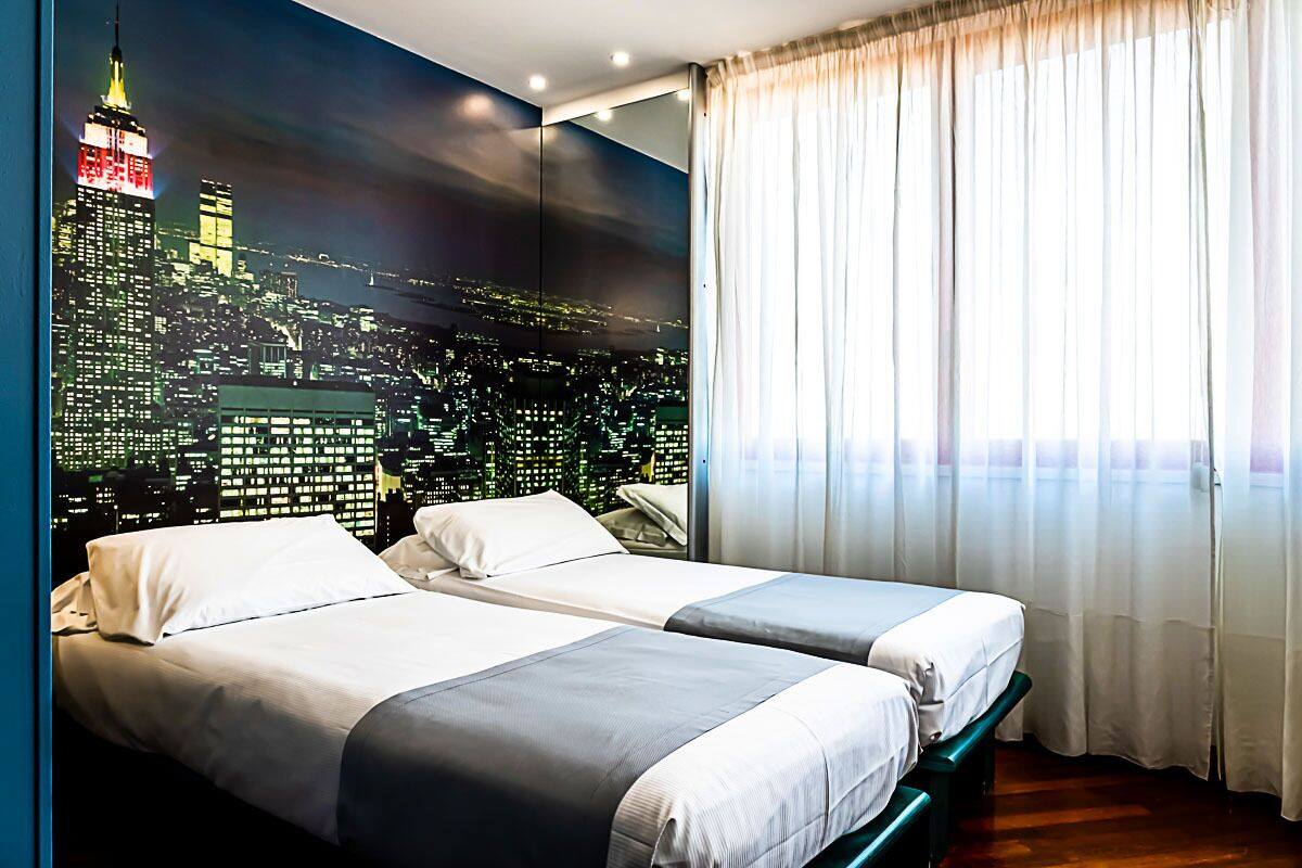 Une chambre d’hôtel avec deux lits, une fresque représentant un paysage urbain sur le mur et une grande fenêtre avec des rideaux.