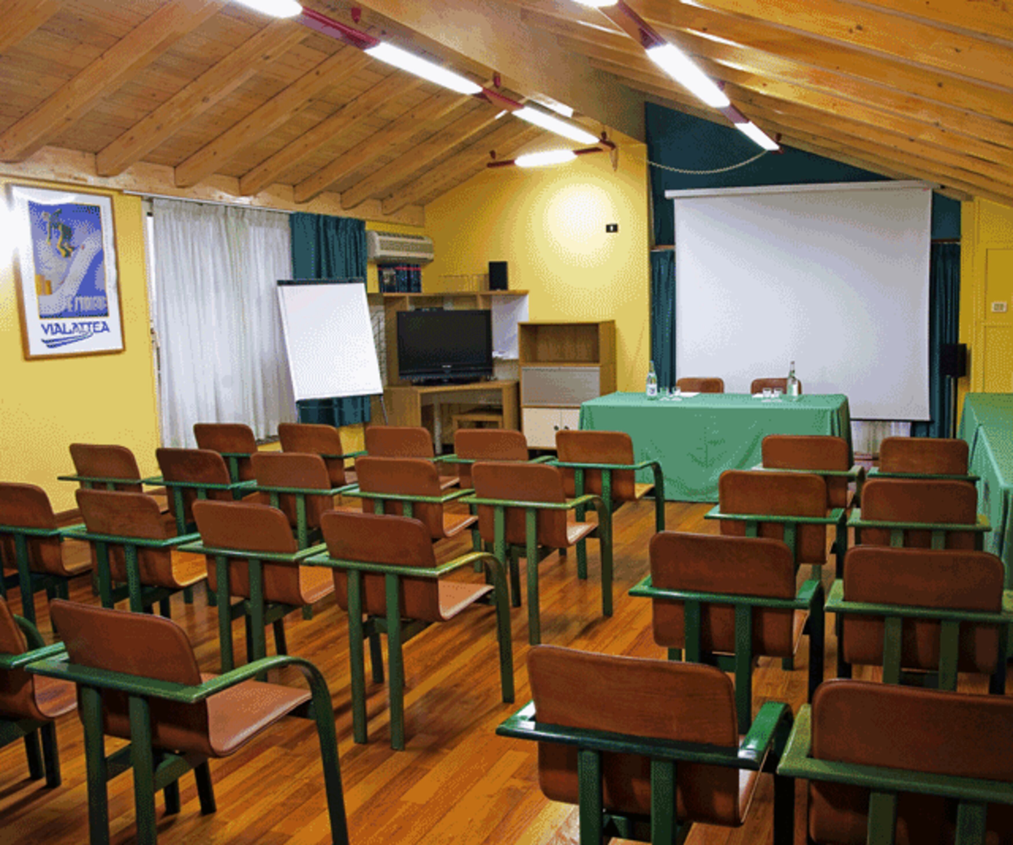 Une salle de classe avec des rangées de bureaux et de chaises, un écran de projection, un tableau blanc et un téléviseur.