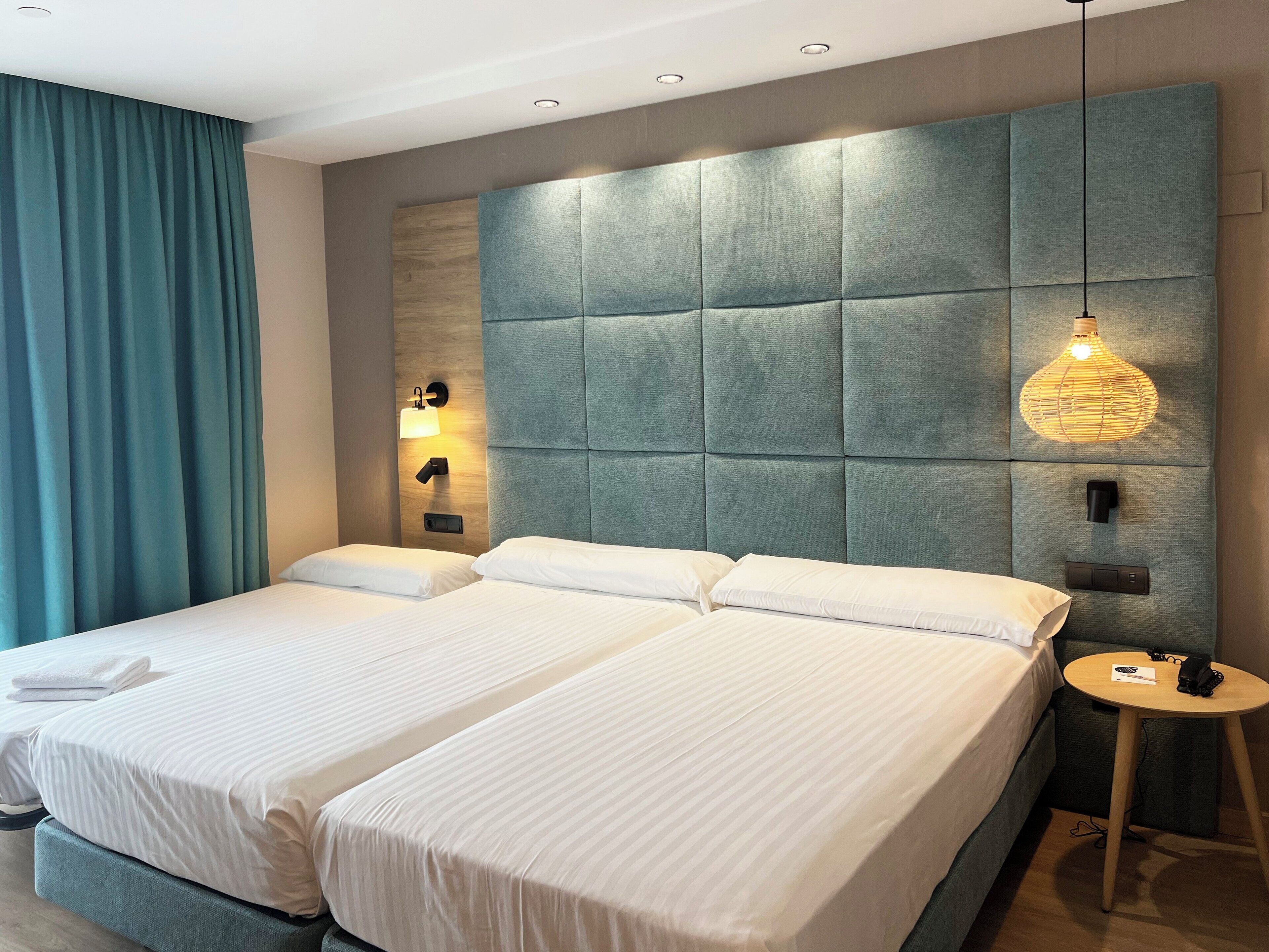 A hotel room with two beds, a large padded headboard, a bedside table with a lamp, and a small round table with a phone and a book.