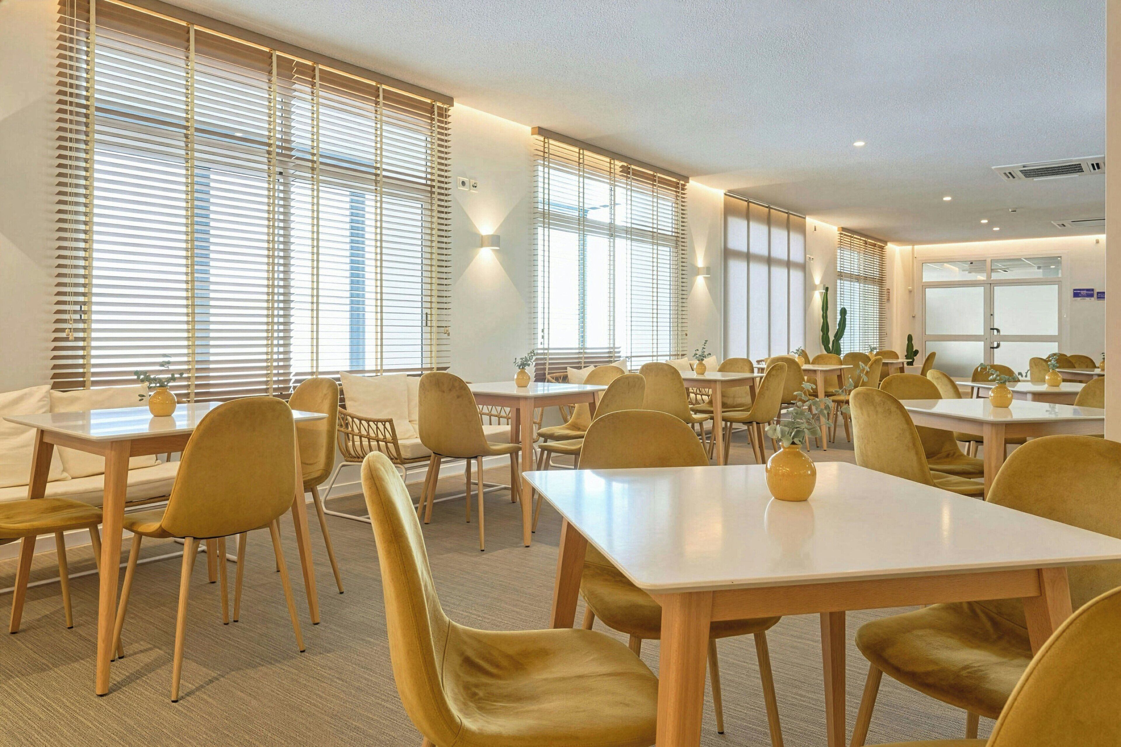 A hotel dining area with multiple tables and chairs, large windows with blinds, and wall-mounted lighting.