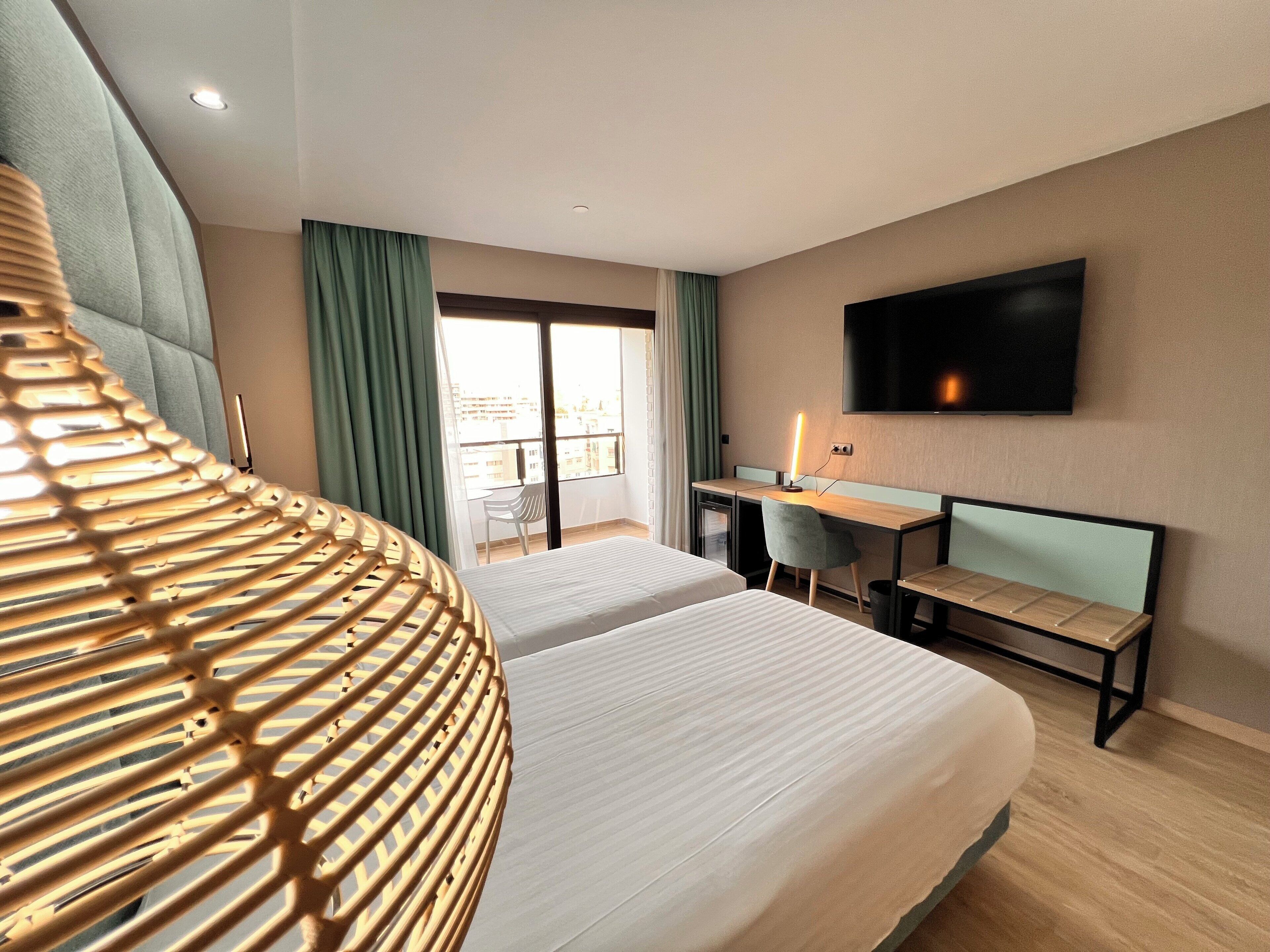 Une chambre d’hôtel avec deux lits, un bureau, une chaise, une télévision et un balcon avec des rideaux.