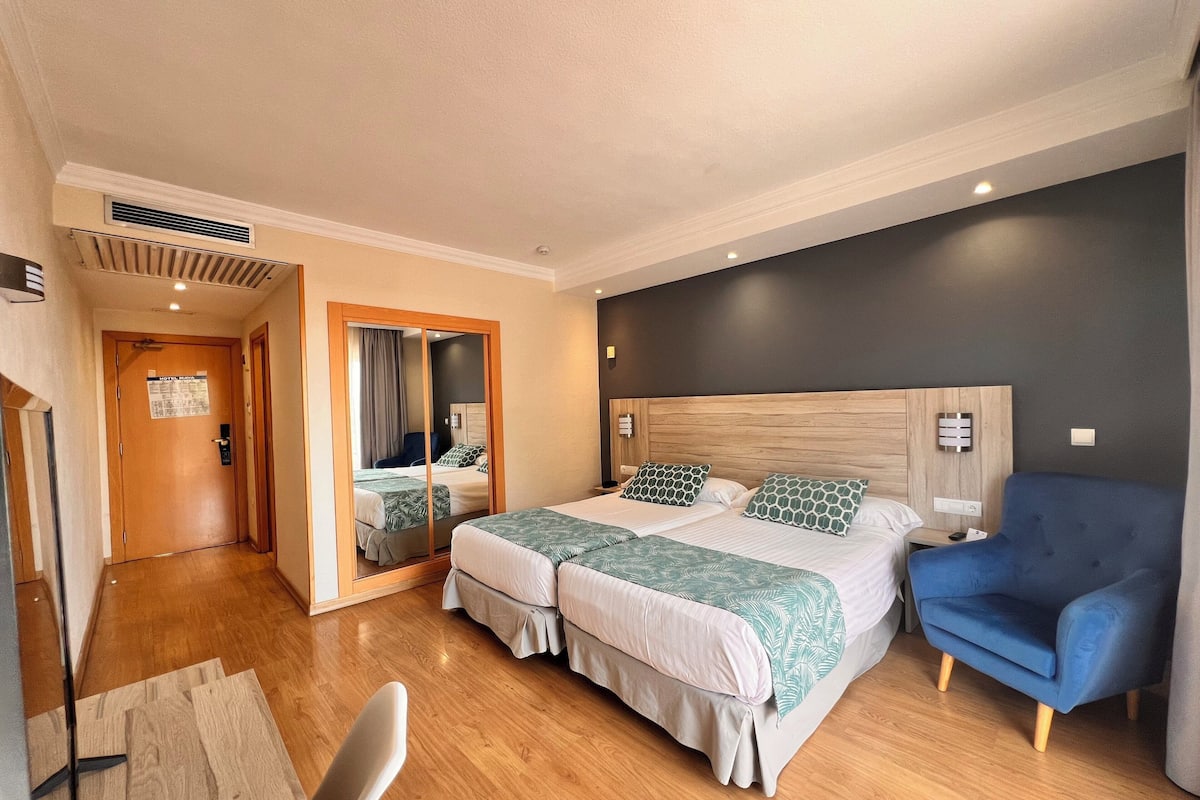 Ein Hotelzimmer mit einem Doppelbett, einem blauen Sessel, einem Holzkopfstück und einem Spiegel.