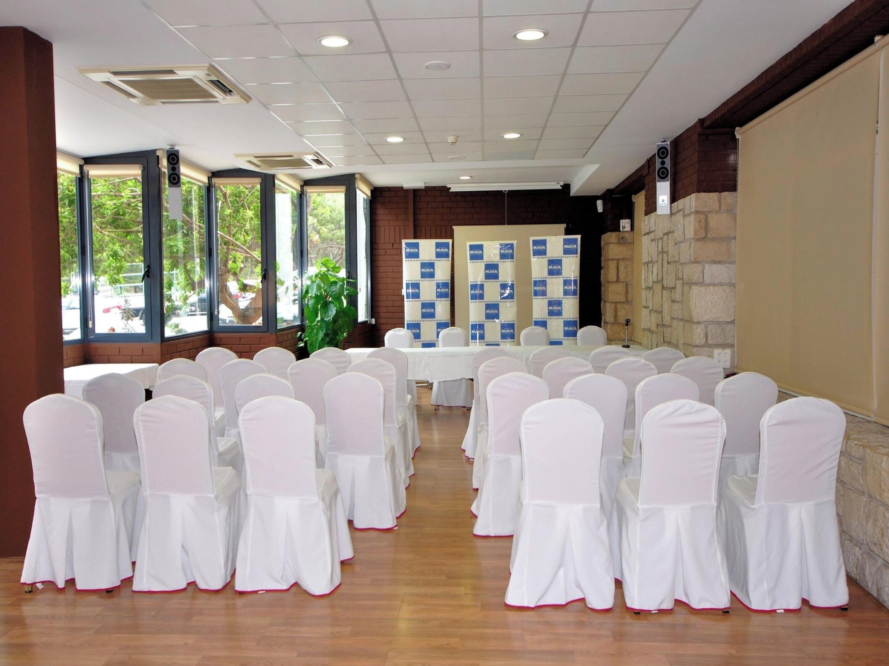 A conference room with white chairs and tables, large windows, and a brick wall.