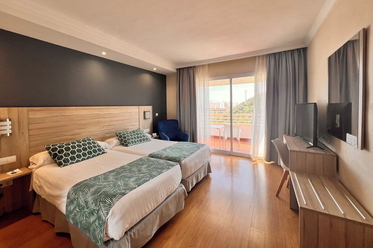 A hotel room with two beds, a desk with a TV, and a balcony with a view.