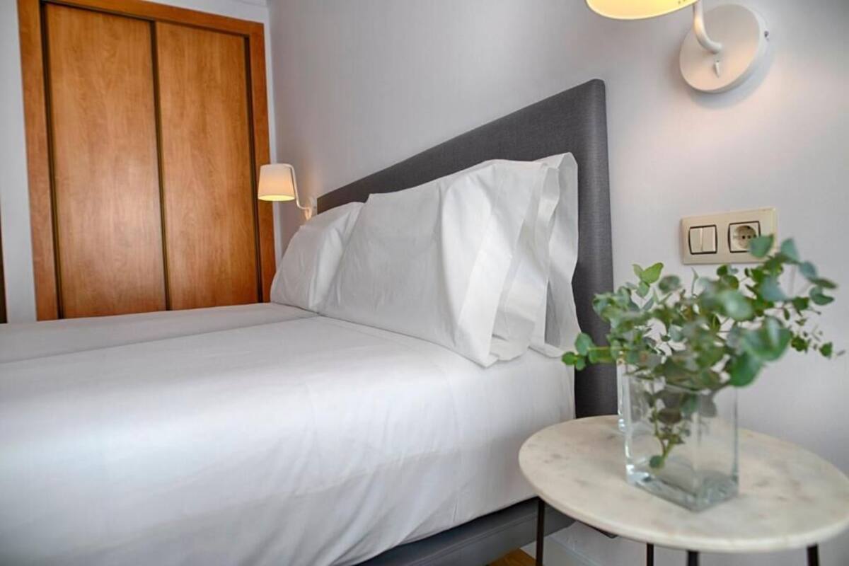 Una cama bien hecha con sábanas blancas, un cabecero gris, una mesita de noche con un florero de plantas verdes y una lámpara fijada a la pared.