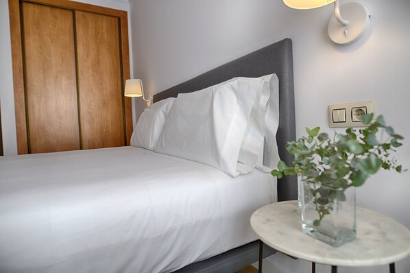 A neatly made bed with white linens, a wooden wardrobe, a bedside table with a vase of green plants, and a wall-mounted light fixture. - Sercotel Tribuna Málaga (Málaga)