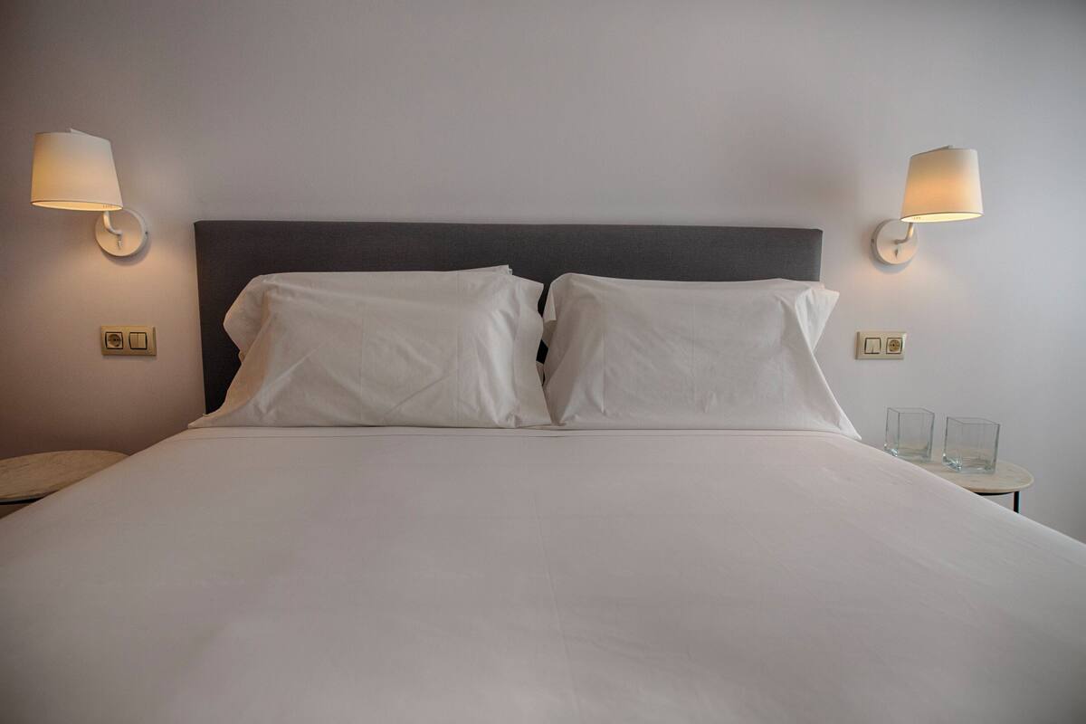 Una cama con sábanas blancas, dos lámparas de pared y una mesita con dos vasos.