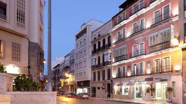 Vista da rua numa cidade, com edifícios, uma rua e carros estacionados.
